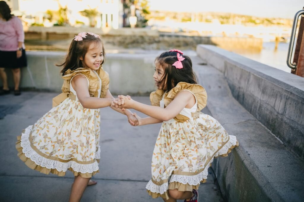 Two twin sisters in floral dresses play joyfully while holding hands outdoors on a sunny day.