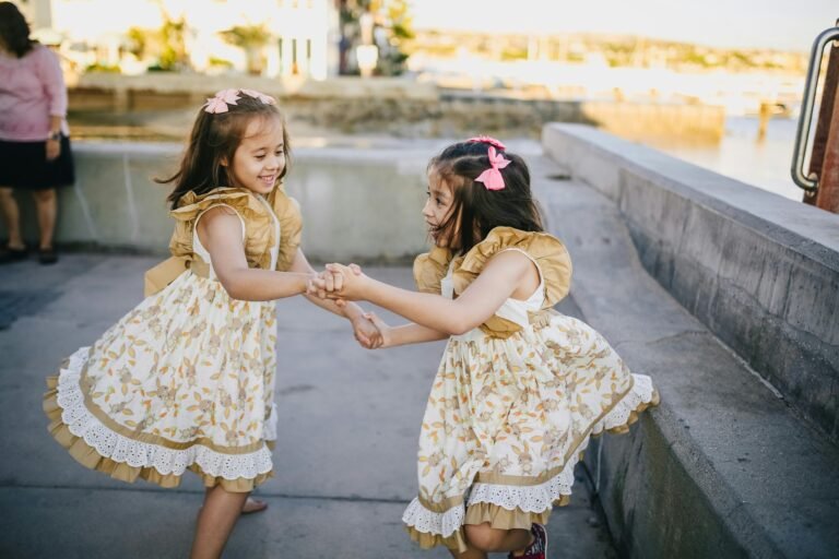 Two twin sisters in floral dresses play joyfully while holding hands outdoors on a sunny day.