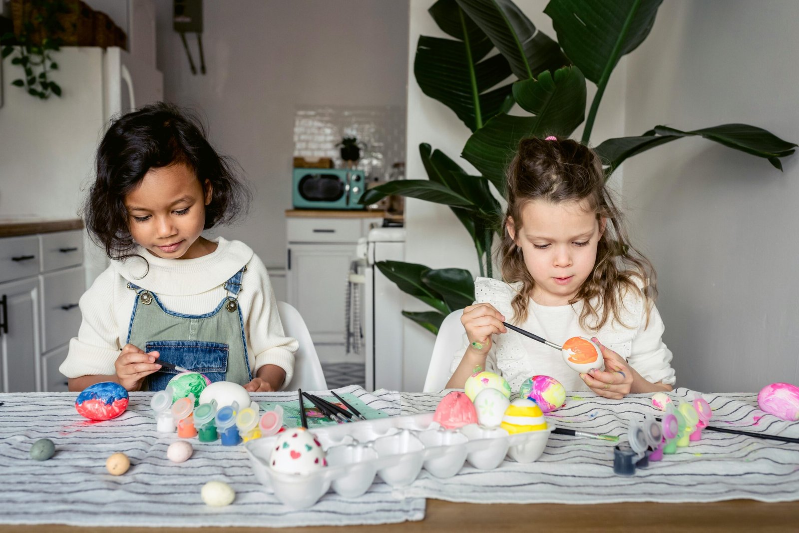 Two children painting colorful Easter eggs at a kitchen table.